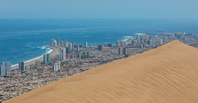 View of the Dragon Dune in the foreground and the city of Iquique in the background, Tarapacá Region, northern Chile. The dune is about 20 000 years old and was originated by coastal winds when the sea level was 100 metres (110 yd) further inside. The dune looks menacing, especially from the bottom, but is stable. It became a Natural Sanctuary in 2005 but before that it was partially removed in the West wing to allow the growth of the city of Iquique. The dune is 6.4 kilometres (4.0 mi) long, between 150–550 metres (160–600 yd) wide and 320 metres (350 yd) high. Iquique has aprox. 185 000 inhabitants and is a prosperous and fast-growing city thanks to the free trade activities.